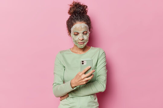 Young beautiful woman with dark curly hair gathered in bun applies nourishing sheet mask takes care of facial skin undergoes beauty treatments wears casual green jumper holds mobile phone chats online