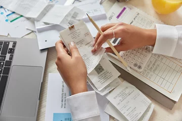 Unrecognizable woman holds receipts pencil uses laptop plans budget manages expenses calculates financial reports checks internet banking service poses at desktop with papers around. View from above © WHstudio Leushin N