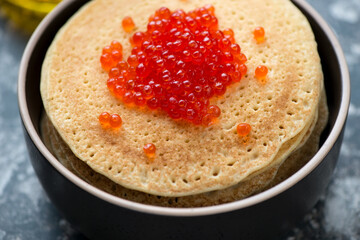 Stack of pancakes served with red caviar in a black bowl, horizontal shot, selective focus, close-up