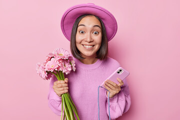 Cheerful dark skinned Asian woman wears elegant hat and knitted jumper holds bouquet of beautiful gerbera flowers and smartphone smiles broadly enjoys festive event isolated over pink background.