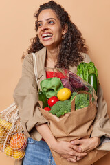Overjoyed Afro American woman carries paper bag full of groceries bought organic vegetables and...