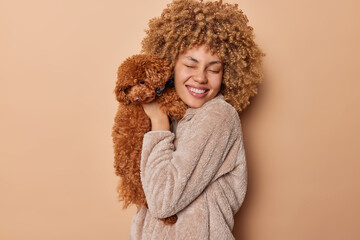 Love between dog and owner. Cheerful woman with curly hair poses with small poodle puppy smiles sincerely dressed in winter coat happy to have best friend isolated over brown studio background
