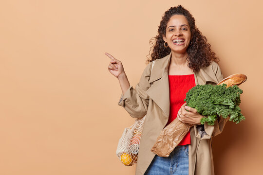 Horizontal shot of happy young woman with Afro hair poses with grocery shopping bag bought fresh products points index finger aside shows direction to supermarket isolated over brown background