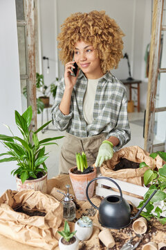 Vertical shot of curly haired skilled female florist involved in process of transplantation makes phone call has satisfied expression surrounded by pots and paper bags with soil stands indoor