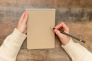 Women's hands in a jumper lie on the table and hold a notebook and a pencil in their hands.