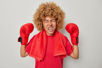 Emotional female boxer with curly hair keeps arms raised wears protective boxing gloves and mouth...