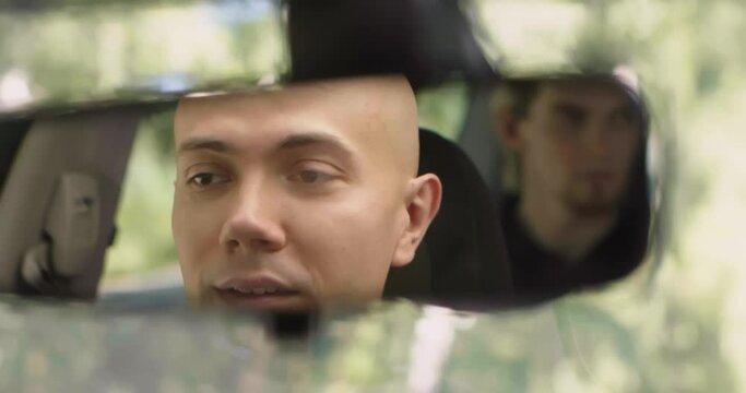 Young man in rearview mirror of car. Stock. Man and friend are driving in car in summer. Bandit-looking men ride in car