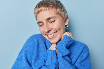 Happy sincere woman with short fair hair smiles broadly keeps eyes closed enjoys pleasant memories wears casual oversized jumper isolated over blue background. Positive human emotions and feelings