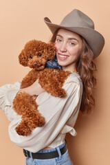 Pretty European female pet owner dressed in beige blouse jeans and cowboy hat poses with poodle dog enjoys company of favorite domestic animal isolated over brown background. Pet care concept