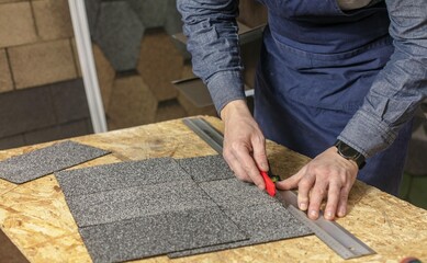 The process of cutting bituminous tiles indoors by a man.
 Close-up of hands and table with tiles and tools. Bituminous tiles on table and walls around. The concept of selling bitumen roof tiles. Buil