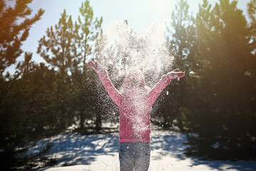 Portrait of a young girl with blonde hair, dressed in bright pink winter clothes and jeans, enjoying the falling snow in a pine forest.The silhouette of a girl who throws snow with her arms wide apart
