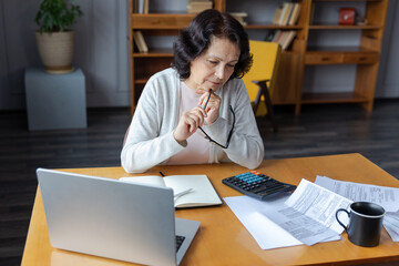 Middle aged senior woman sit with laptop and paper document, Pensive older mature lady reading paper bill pay online at home managing bank finances calculating taxes planning loan debt pension payment