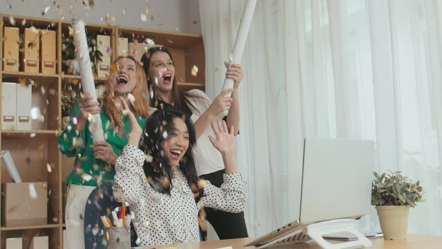 Two cheerful female employees exploding flappers with confetti while Asian businesswoman clapping hands by workplace in office and looking at laptop screen