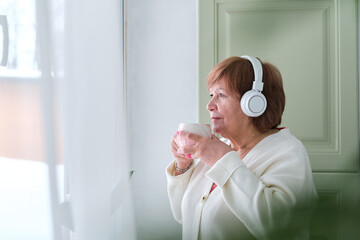 elderly woman standing by a window, lost in her thoughts while listening to music or a podcast, to...
