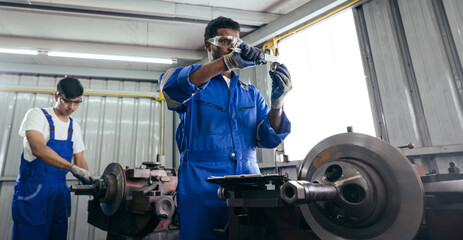 African American mechanic worker inspecting on equipment part while colleague working on lathe...