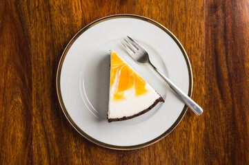 Piece of cheesecake with orange mousse on a white plate with a golden edge on a wooden table.