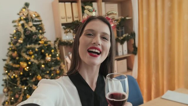 Happy young woman in xmas headband wishing young merry Christmas and drinking red wine while communicating in video chat against decorated firtree