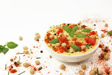 Hummus with tomatoes and paprika on a white background in a plate topped with chickpeas and sesame seeds, basil and spices. Culinary and food menu photo. Eastern or Mediterranean cuisine.