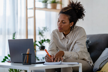 Young African Ethnicity Freelancer Woman Working On Laptop At Home Office And Taking Notes, Sitting At Desk Near Window, Looking At Screen, Writing Down Information From Computer, Enjoying Remote Work