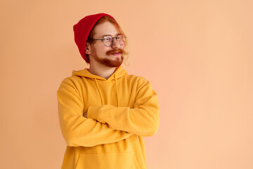 A stylish red-haired handsome man in glasses and a red cap and a yellow hoodie is looking away and smiling. Copy space.