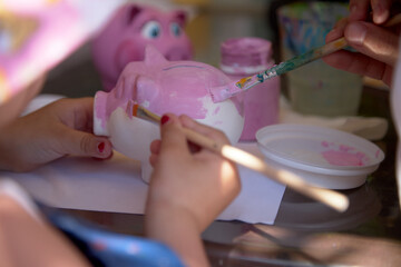 The child's hand holds a brush and paints a clay piggy bank in the shape of a pig