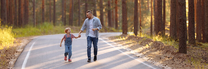 Dad and his little daughter are walking along a forest road among tall pines. Family walk in the forest at sunset, man and little girl. Banner for website header design with copy space.