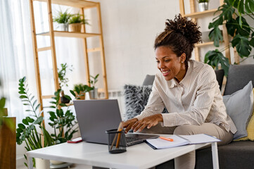 Young African Ethnicity Freelancer Woman Working On Laptop At Home Office And Taking Notes, Sitting At Desk Near Window, Looking At Screen, Writing Down Information From Computer, Enjoying Remote Work