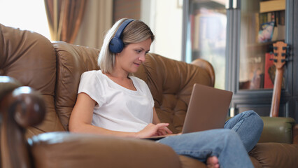 Female freelancer with headphones and laptop works online while sitting on couch.