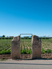 Una puerta moderna y minimalista formada por dos antiguos pilares de piedra sirve de entrada a un gran campo verde cultivado bajo un cielo azul claro.