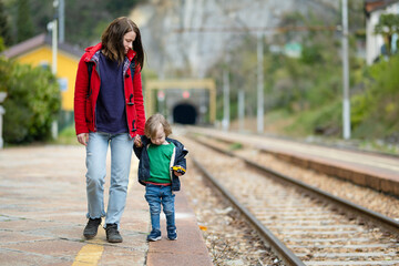 Young mother and her toddler son on a railway station. Mom and little child waiting for a train on a platform. Family ready to travel. Going on vacation with kids.