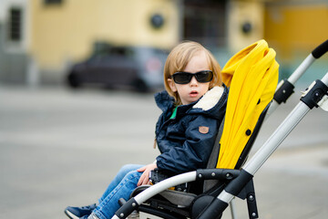 Naklejka premium Sweet toddler boy sitting in a stroller outdoors. Little child in pram. Infant kid in pushchair. City walks with kids. Family leisure with a child.