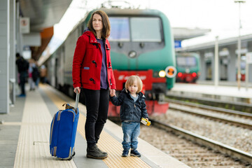 Young mother and her toddler son on a railway station. Mom and little child waiting for a train on a platform. Family with a luggage suitcase ready to travel. Going on vacation with kids.