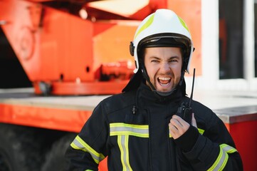 Fireman in a protective uniform standing next to a fire truck and talking on the radio.