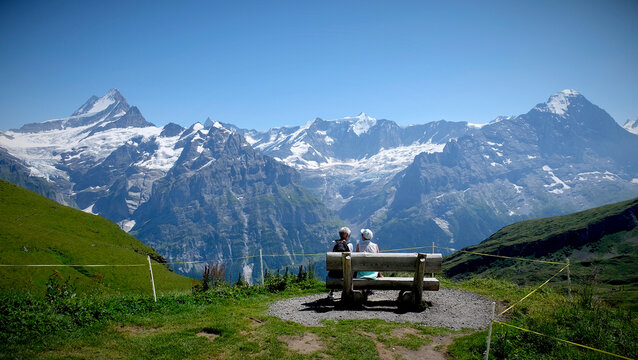 Elderly leadies sat on a wooden bench chat while watching the snowy peaks of Grindelwald region in Switzerland. Summer panorama with deep blue sky