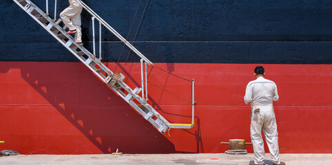 Two Engineers are walking up the white gangway accommodation ladder of red and black Oil Tanker to...