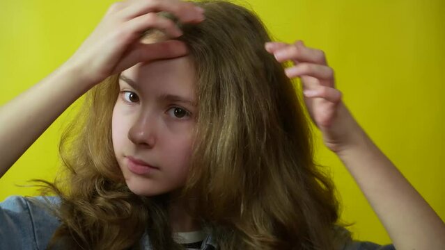 Close-up view of adolescent girl looking to the camera while touching her curly hair standing isolated over yellow background