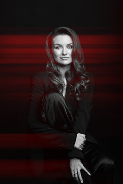 Woman portrait with black suit and big wavy hair in red color split effect style. Model sitting on chair and hugging her one leg. Woman looking at camera. Futuristic looking style