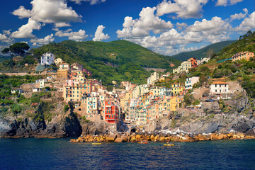 Fototapeta na wymiar Picturesque village of Riomaggiore against a blue sky with white clouds. View from the sea side of the colourful houses, green terraced vineyards, tourists enjoying holidays. Cinque Terre, Italy.