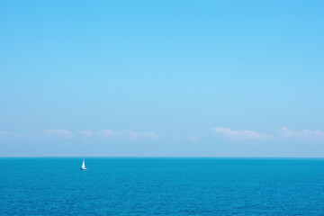 Fototapeta na wymiar Tiny sailboat in the distance on the empty sea with empty sky and tiny clouds 
