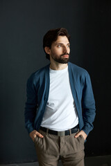 Obraz premium Image of fashionable brutal handsome man with beard and stylish hairstyle standing against black studio wall in blue cardigan and plaid brown pants, looking aside with hands in pockets
