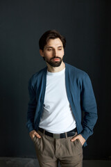 Fototapeta premium Portrait of handsome brutal man teacher in cardigan and stylish pants standing against black studio background with hands in pockets, looking down, preparing his mind for productive work day