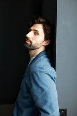 Clever gorgeous tempting attractive hot brutal bearded guy standing against black background leaning his back on gray wall, looking at you turning his head to camera, trying to attract your attention