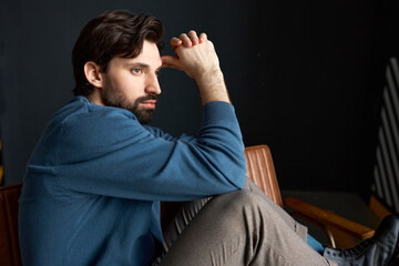 Indoor portrait of young manly guy with beard in cardigan sitting on couch touching his forehead,...