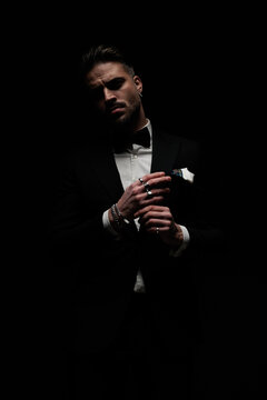 stylish unshaved groom in tuxedo touching fingers and posing