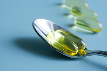 Transparent yellow capsules with fish oil and a silver teaspoon on a blue background. Vitamin D and...