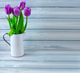 White jug with purple tulips on a wooden light wooden background