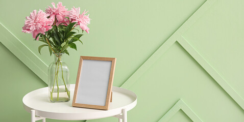 Blank photo frame and vase with bouquet of pink peony flowers near green wall in room. Banner for design