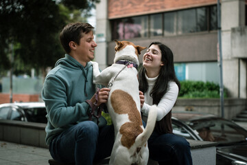 A smiling couple is with their dog in the city. Happy dog with his family in the city. Cars and gardens are out of focus in the background