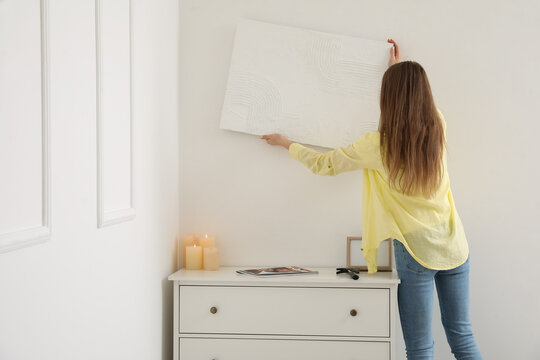 Young woman hanging painting on light wall at home, back view