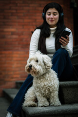 A smiling girl looks at her dog while listening to music on her phone. Relaxed girl with her dog listening to music from the cell phone. Girl with headphones.